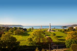 View of Plymouth Sound from the Crowne Plaza Hotel in Plymouth. The foreground is a big green park with a tall memorial monument standing, and a red and white striped lighthouse standing slightly further to towards the waterfront.